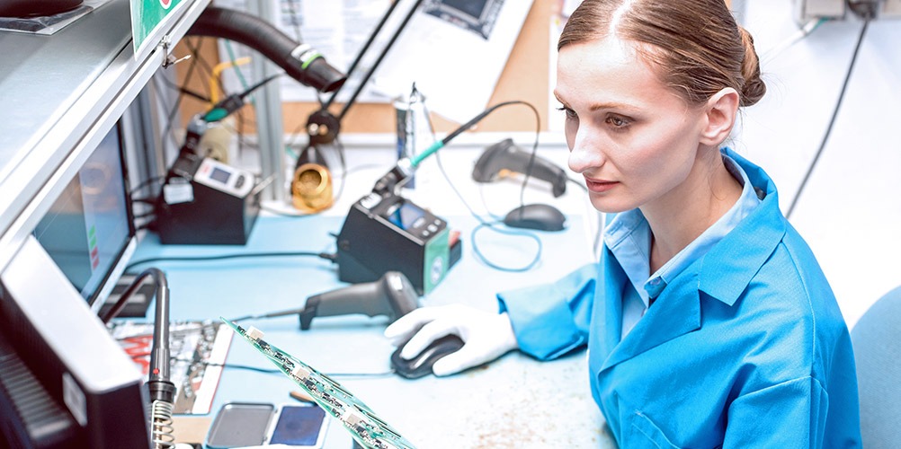 A supply chain technician looks at a computer screen while holding a processor board